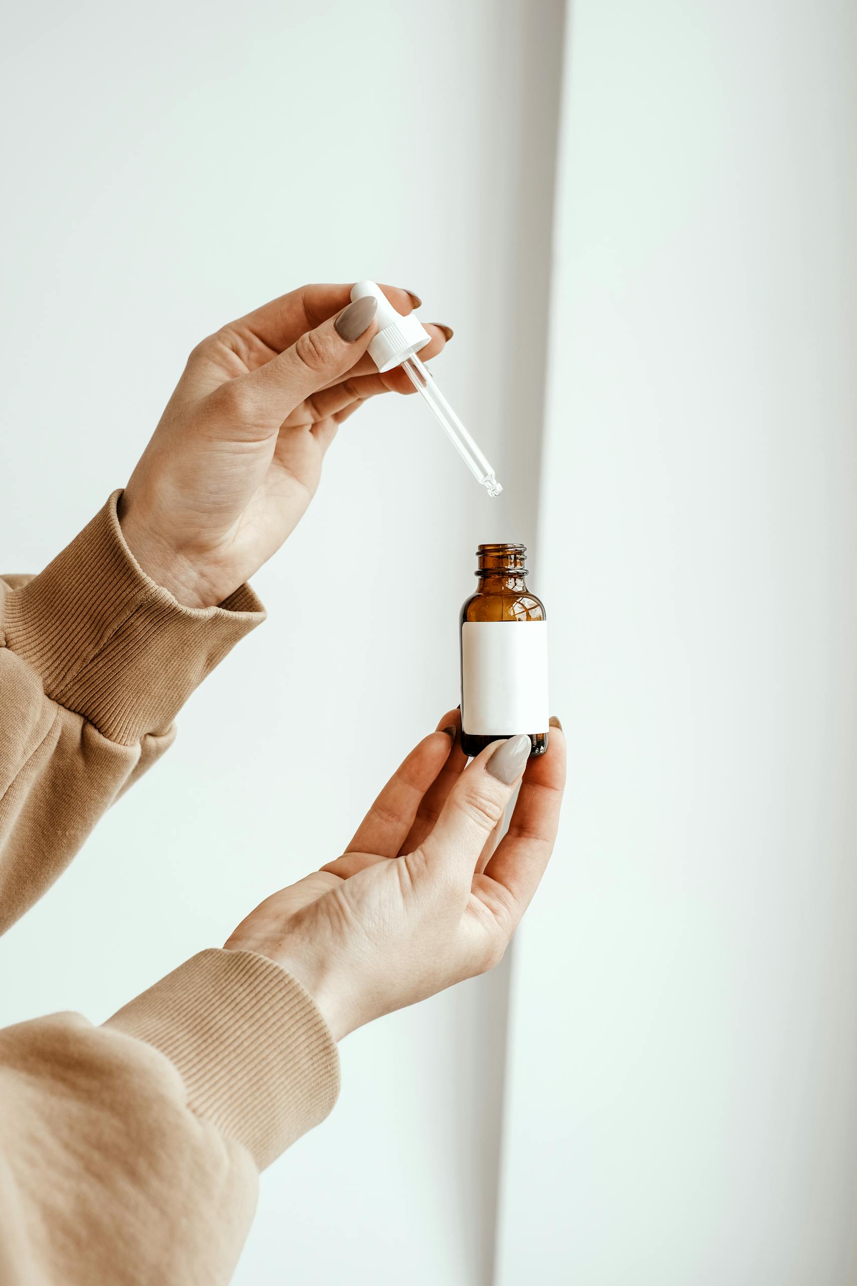 A close-up of hands holding a skincare dropper bottle with a beige sweater and blank label.