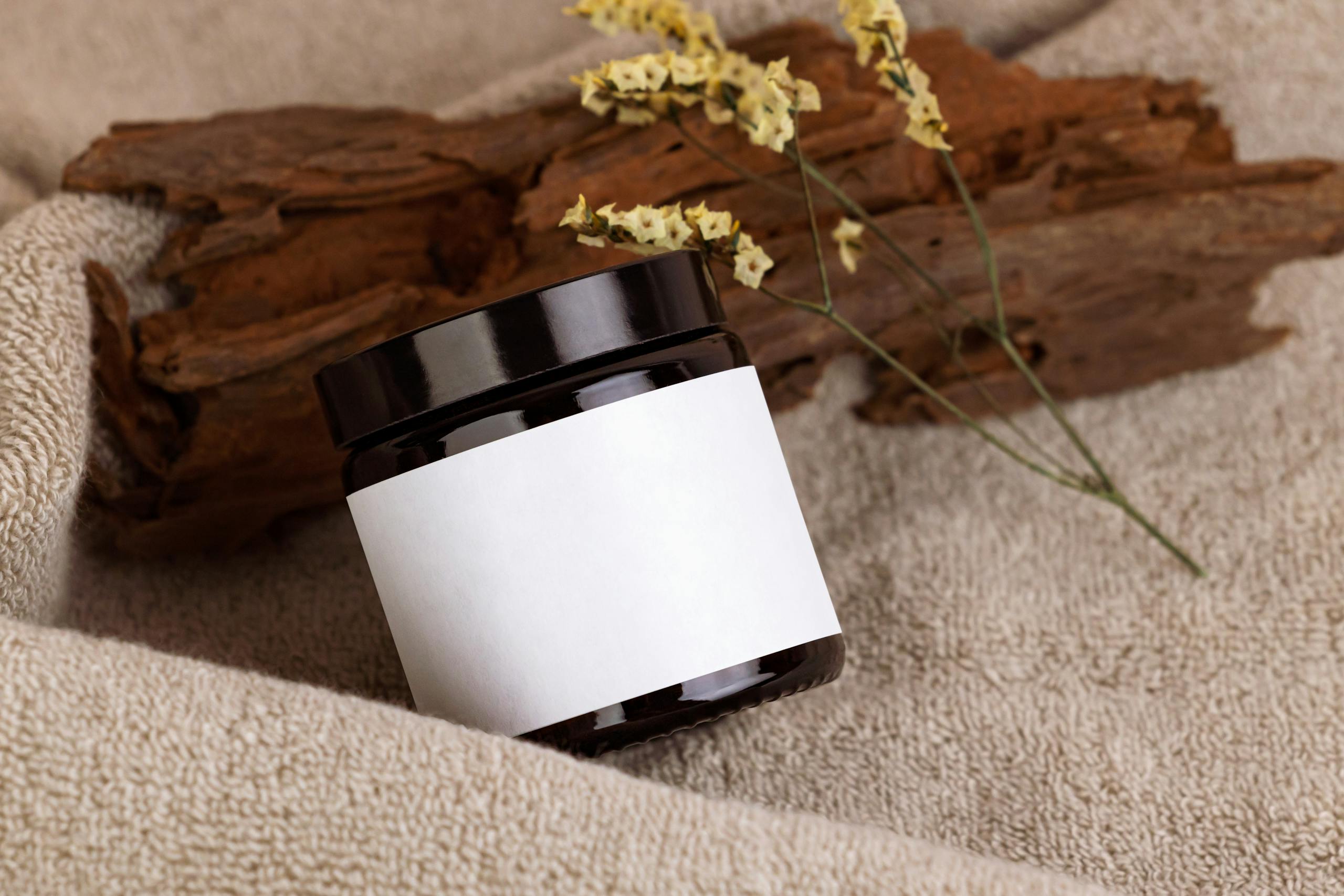 A minimalist black cosmetic jar with blank label on a natural textured towel background, accented by dried flowers and wood.