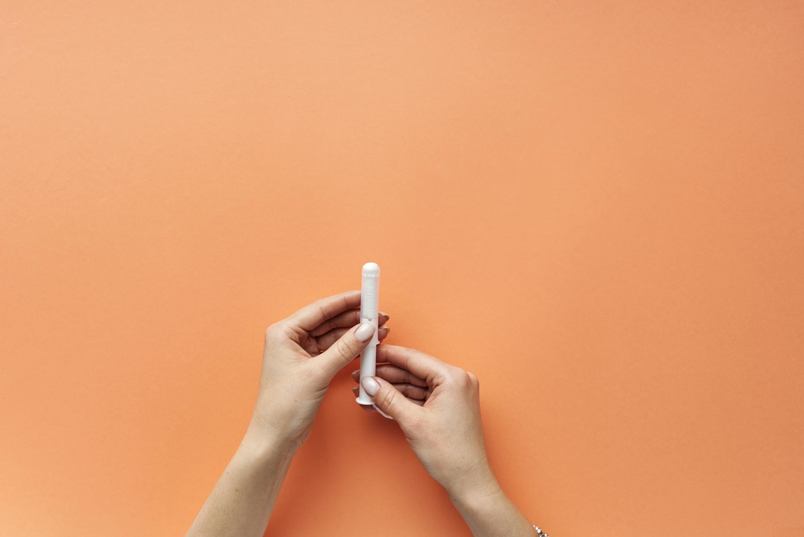 Close-up of female hands holding a tampon on an orange background, symbolizing menstrual hygiene.